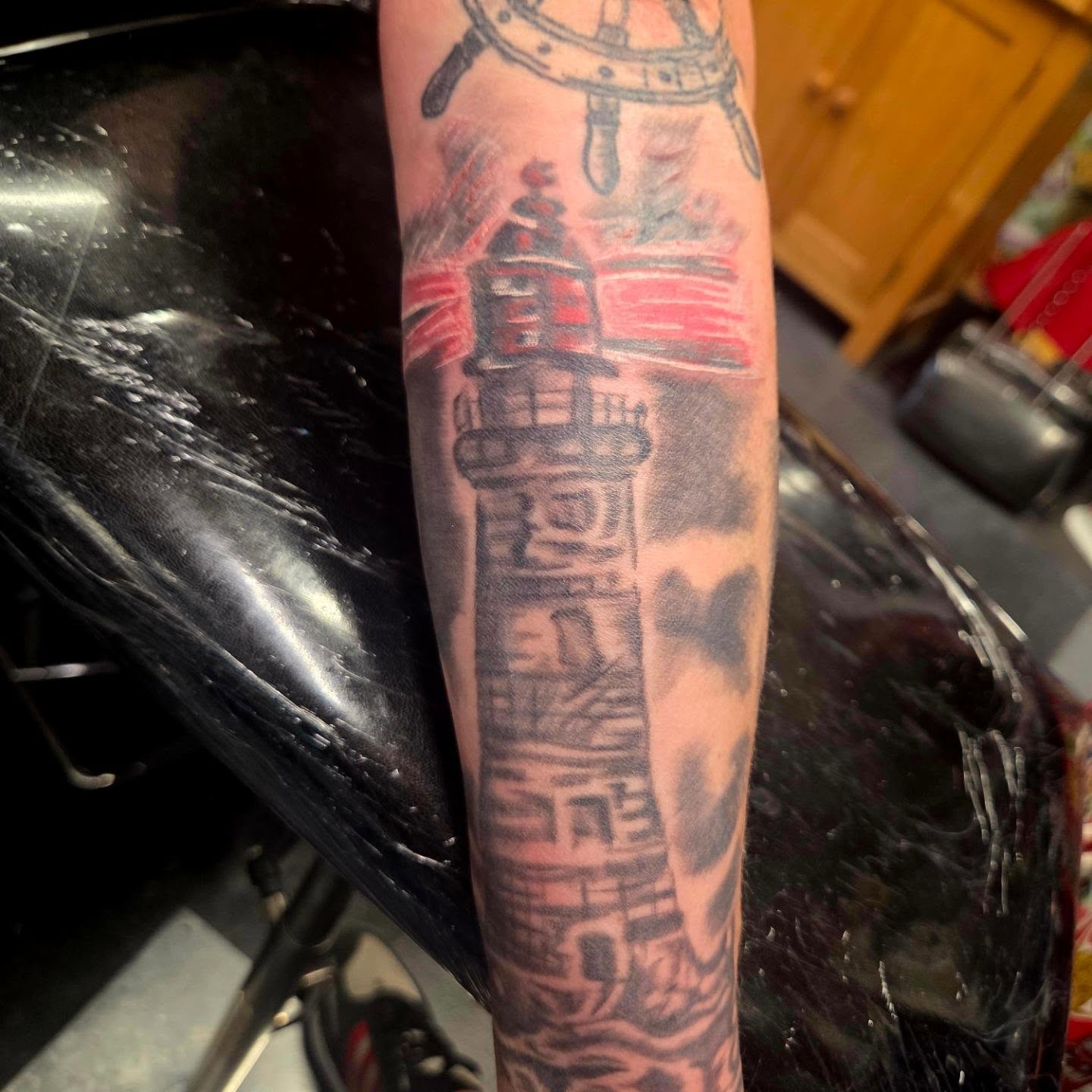 Lighthouse scene tattoo with black shading and red accents on a forearm